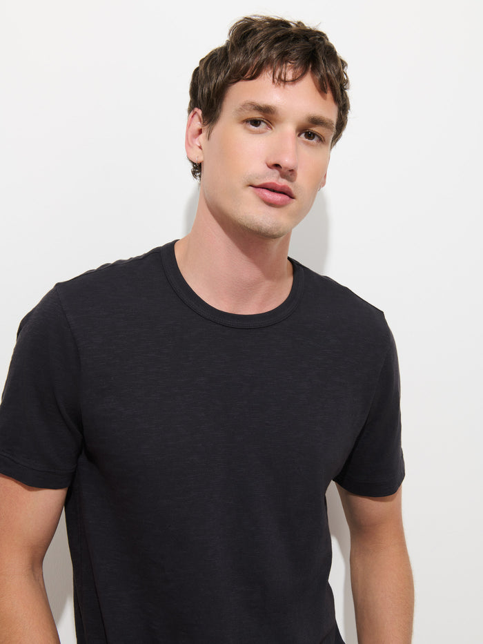 A man with short brown hair stands against a white background, wearing The Standard T-Shirt In Slub Cotton—a plain black men’s tee crafted from breathable fabric—while looking at the camera with a neutral expression.