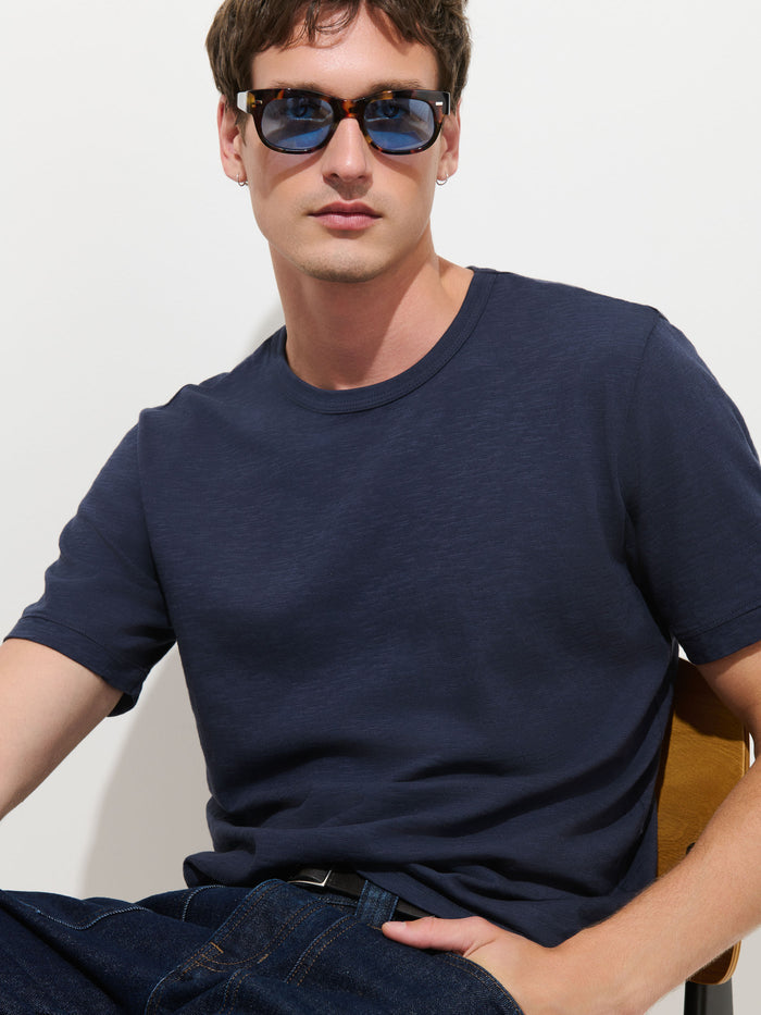 A man wearing blue sunglasses, The Standard T-Shirt In Slub Cotton in navy, and dark jeans sits on a chair against a white background, looking at the camera with a relaxed expression.