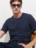 A man wearing blue sunglasses, The Standard T-Shirt In Slub Cotton in navy, and dark jeans sits on a chair against a white background, looking at the camera with a relaxed expression.
