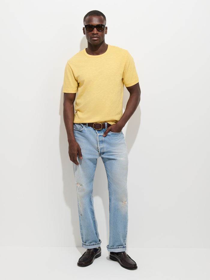 A man wearing sunglasses, The Standard T-Shirt In Slub Cotton in yellow, light blue distressed jeans, a brown belt, and dark shoes stands against a plain white background.