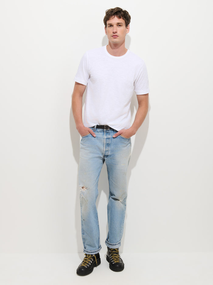 A person stands against a white background wearing The Standard T-Shirt In Slub Cotton—a plain white men’s tee made from breathable slub cotton—paired with light blue ripped jeans and black boots, hands tucked in pockets.