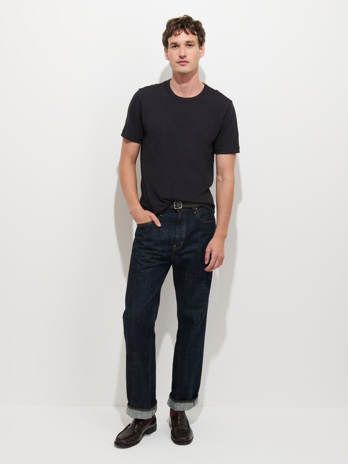A man stands against a white background, wearing The Standard T-Shirt In Slub Cotton—a black men’s tee crafted from breathable fabric—paired with dark cuffed jeans, a belt, and black shoes. One hand rests in his pocket as he maintains a neutral expression.