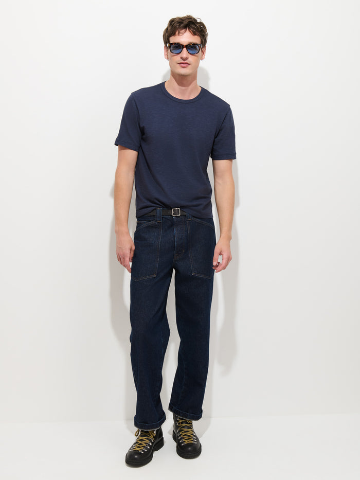 A man wearing sunglasses, The Standard T-Shirt In Slub Cotton—a navy blue men’s tee made from breathable fabric—dark blue jeans, and black boots with yellow laces stands against a plain white background.