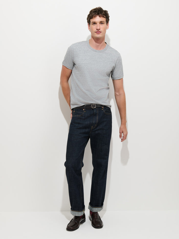 A man stands against a white background, wearing the Standard T-Shirt In Slub Cotton—a light gray men’s tee crafted from breathable fabric—paired with dark blue cuffed jeans and dark brown shoes.