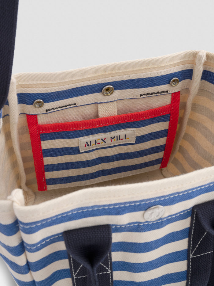 The Striped Mini is an open tote bag made of striped cotton canvas in blue and beige, featuring a red-outlined inner pocket and a sewn-on ALEX MILL label.