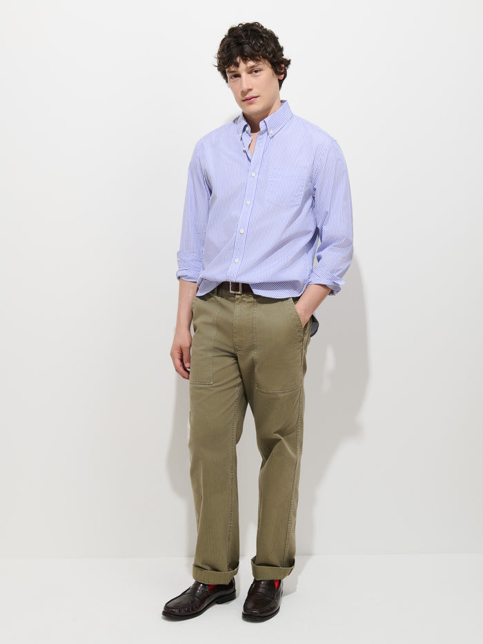 A person stands against a plain white background, wearing the Mill Striped Shirt in Thomas Mason—a light blue striped button-down—paired with olive green pants and dark brown shoes, with one hand in their pocket.