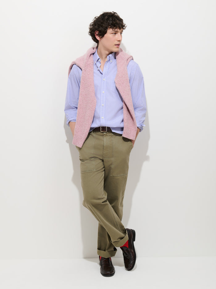 A person with curly hair wears the Mill Striped Shirt in Thomas Mason, paired with olive pants, dark shoes, and a pink sweater draped over their shoulders. They are standing against a white background with arms relaxed and legs crossed.