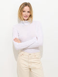 A woman with blonde hair stands against a plain white background, smiling with her arms crossed while wearing a Tissue Turtleneck In Cotton and light beige pants.