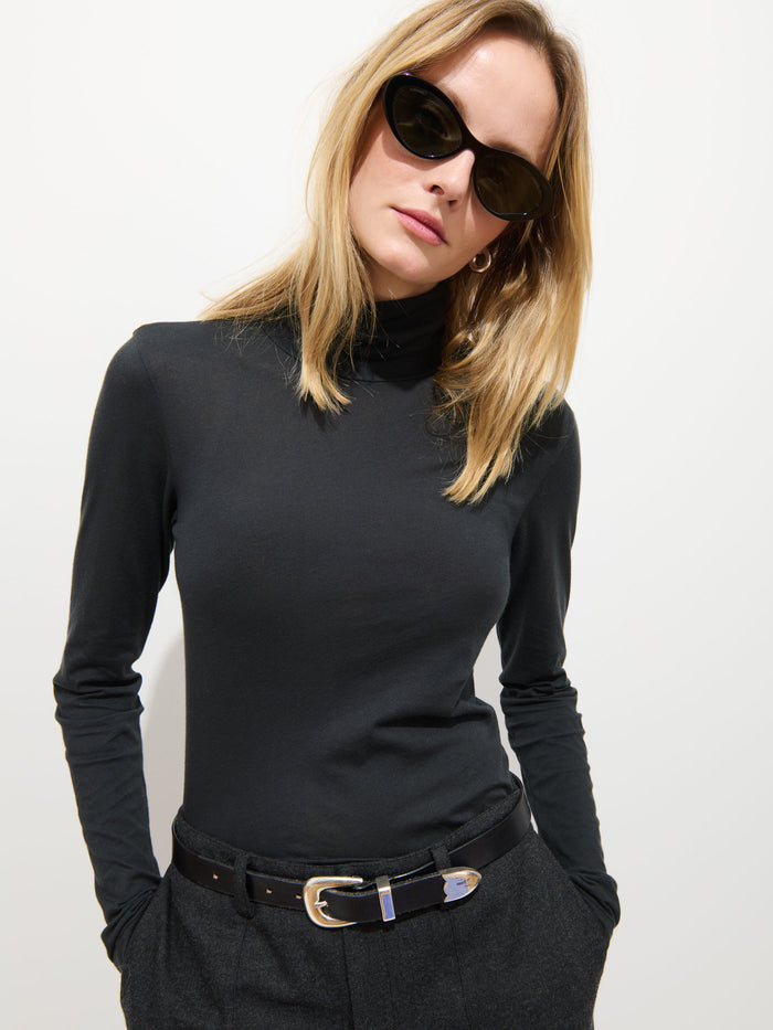 A woman with straight blonde hair wears black cat-eye sunglasses, the Tissue Turtleneck In Cotton, and dark trousers with a black belt; she stands against a plain white background.