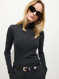 A woman with straight blonde hair wears black cat-eye sunglasses, the Tissue Turtleneck In Cotton, and dark trousers with a black belt; she stands against a plain white background.