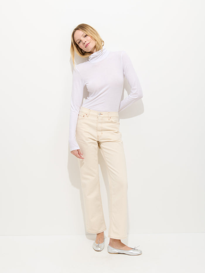 A woman stands against a white background, wearing the Tissue Turtleneck In Cotton, cream pants, and silver ballet flats. She leans slightly to one side with her head tilted and arms relaxed by her sides.