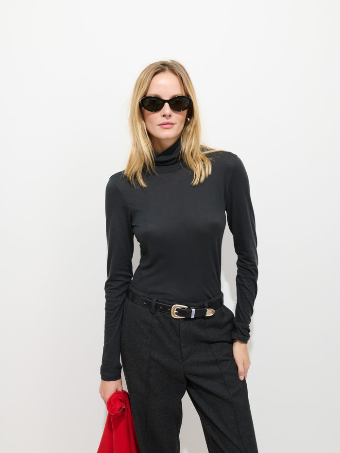 A woman with blonde hair and a confident expression stands against a white background, wearing the Tissue Turtleneck In Cotton in black, dark trousers, and sunglasses. She holds a red garment in one hand.