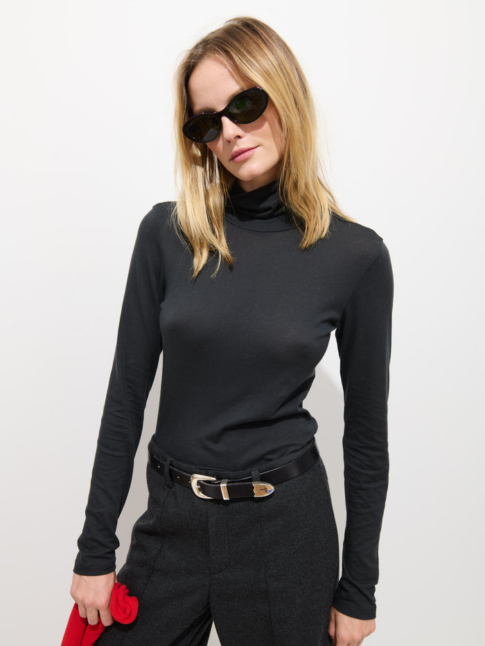 A woman in dark sunglasses, a black Tissue Turtleneck In Cotton, black belt, and dark pants stands against a white background, holding a red item in her left hand.