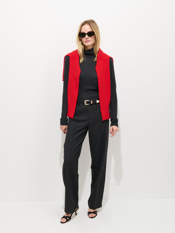 A woman with straight blonde hair and a neutral expression stands against a white background, wearing black sunglasses, black trousers, black sandals, a red sweater over her shoulders, and the Tissue Turtleneck In Cotton.