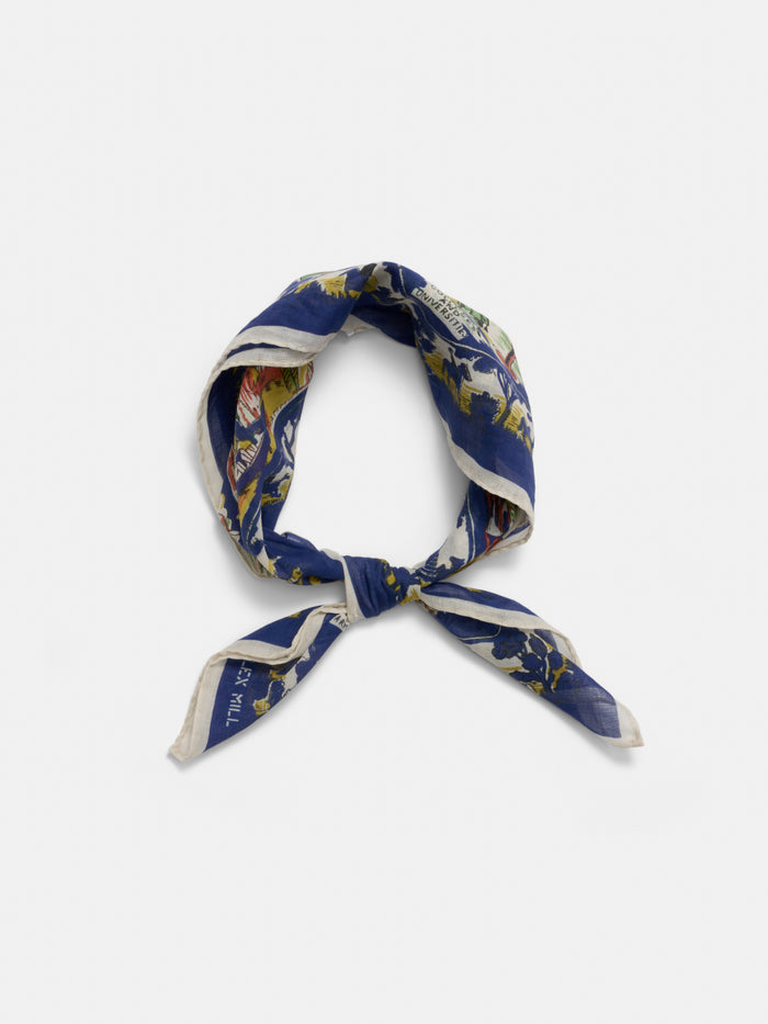 The Travel Scarf In Cotton Voile is a square scarf made from soft cotton voile, showcasing a blue, yellow, and white pattern. It’s styled in a loose knot on a plain white background for a timeless vintage bandana look.