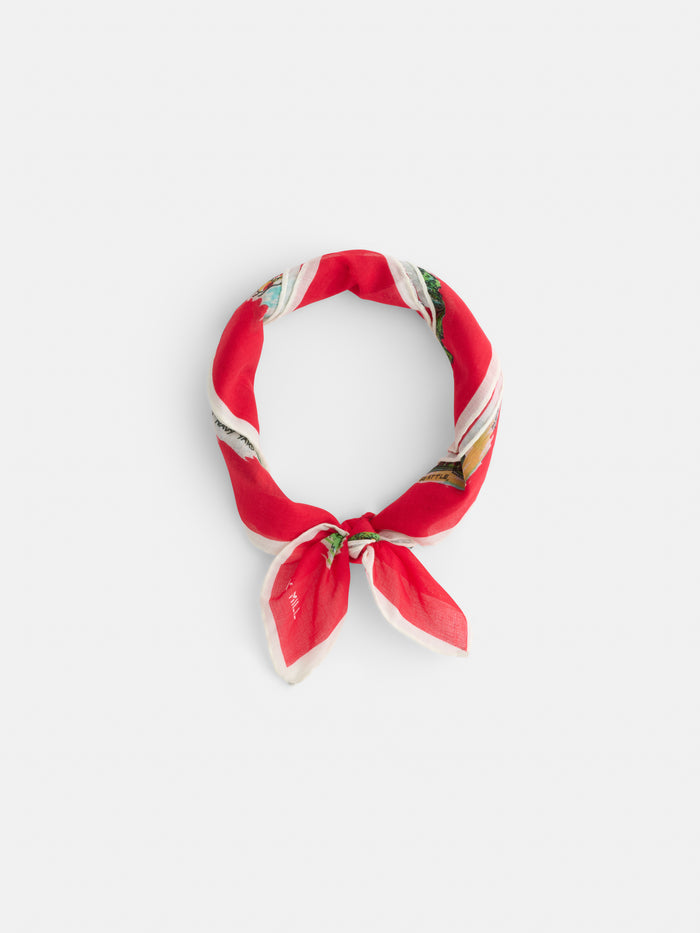 The Travel Scarf In Cotton Voile is a red square scarf with white, light blue, and green accents, styled like a vintage bandana and tied with pointed ends. It’s displayed on a plain white background.