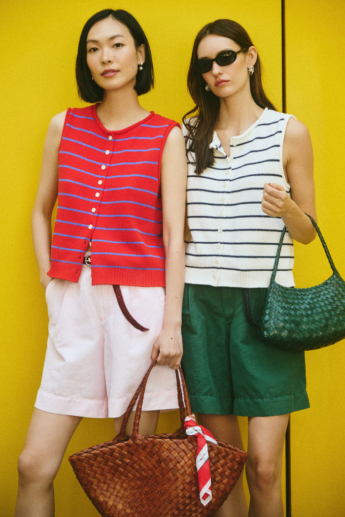 Two women stand by a bright yellow wall with woven bags and playful NYC vibes. One wears airy pink shorts and a vintage-inspired Travel Scarf in Cotton Voile; the other pairs green shorts with a red and white striped sleeveless top.