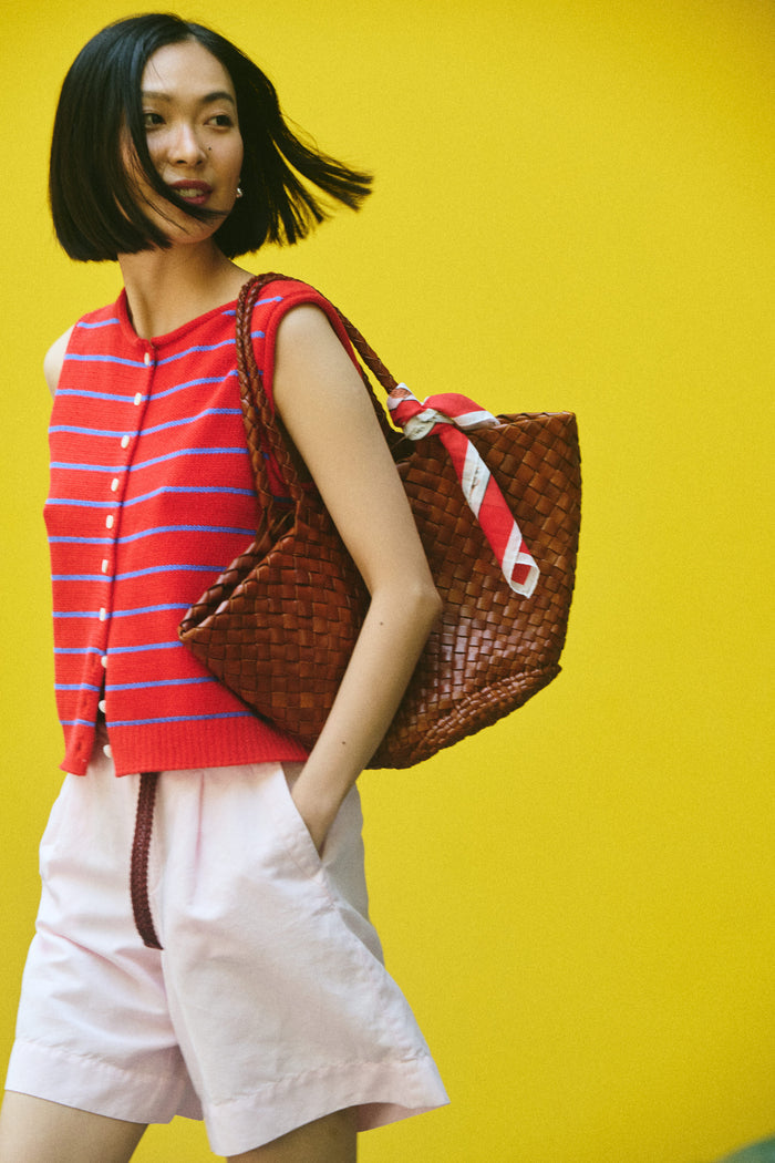 A woman with a bob haircut stands against a bright yellow background, wearing a red and blue striped sleeveless top, light pink shorts, and the Travel Scarf In Cotton Voile as a vintage bandana, carrying a large woven brown bag.