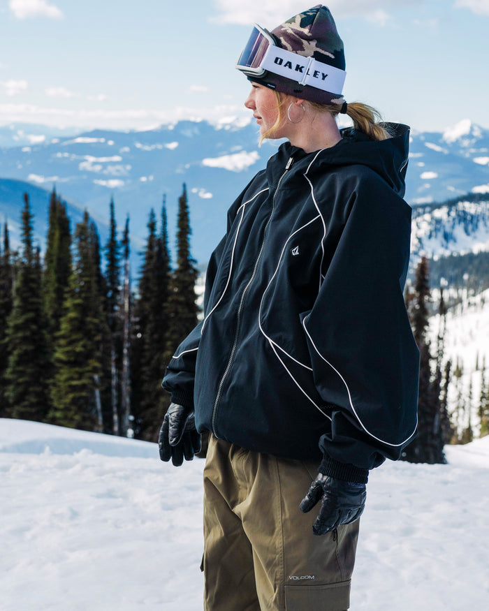 A person in winter gear stands on a snowy mountain, wearing the Volcom Unisex Liftline 20K Jacket, khaki pants, gloves, and an OAKLEY beanie with goggles. Pine trees and mountain peaks are visible in the background.