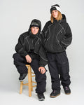 Two people in Volcom Unisex Liftline 20K Jackets, black pants, and winter hats pose together—one standing and one seated on a stool. Both display neutral expressions and are dressed for cold weather in women’s snowboarding apparel, featuring the baggy-fit Volcom jacket designed for style and comfort on the slopes.