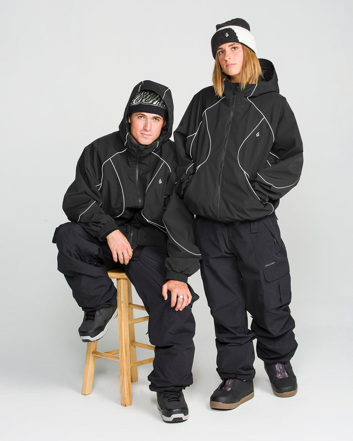 Two people in Volcom Unisex Liftline 20K Jackets, black pants, and winter hats pose together—one standing and one seated on a stool. Both display neutral expressions and are dressed for cold weather in women’s snowboarding apparel, featuring the baggy-fit Volcom jacket designed for style and comfort on the slopes.