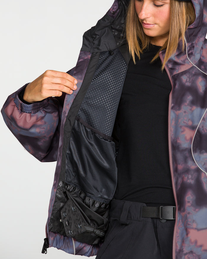 A person models the Volcom Unisex Liftline 20K Jacket, showcasing its purple patterned hooded design and baggy snowboard fit while holding it open to reveal black interior mesh pockets. This 20K waterproof jacket is styled with a black shirt and pants, with only part of their face visible.