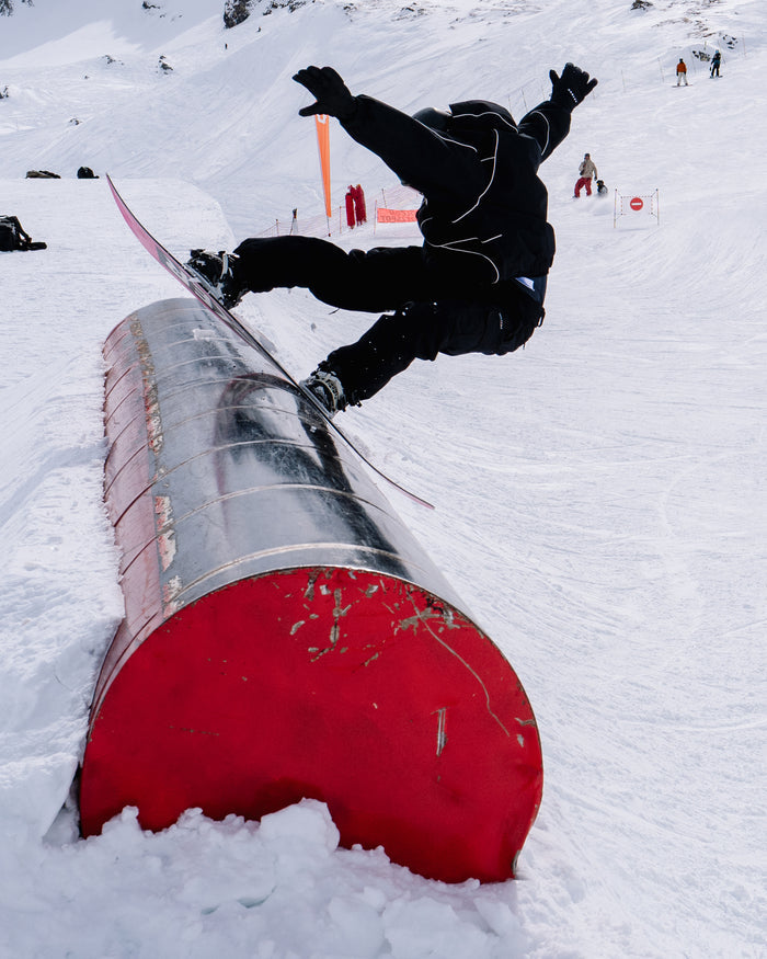 Wearing the Volcom Unisex Liftline 20K Jacket in a baggy fit and black gear, a snowboarder balances mid-trick on a red rail in a snowy terrain park, arms raised, as onlookers watch in the background.