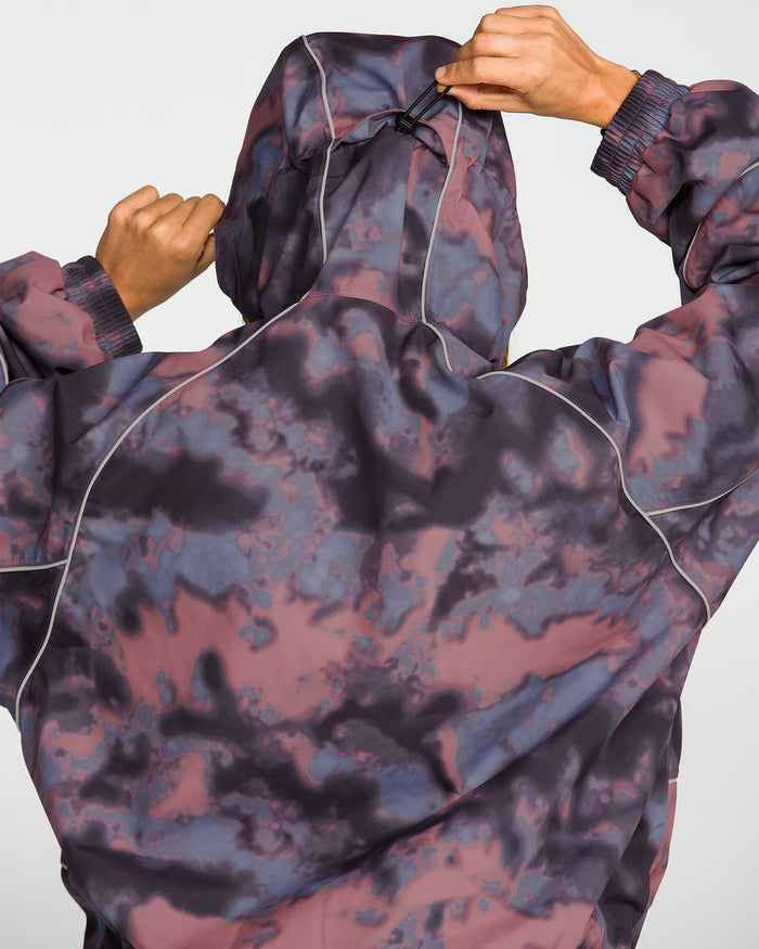 A person, seen from behind, adjusts the hood of a purple and pink marbled Volcom Unisex Liftline 20K Jacket with light piping. Their hands are raised, holding the drawstring at the top of the hood on this baggy fit snowboard jacket.