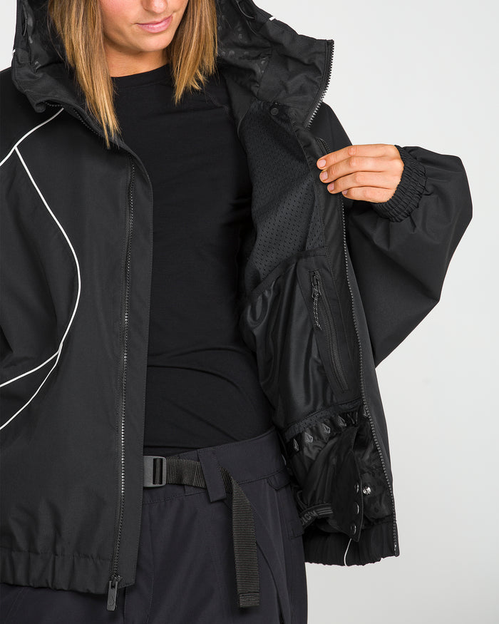 A person is wearing the Volcom Unisex Liftline 20K Jacket in black, featuring a relaxed, baggy fit and a hood. The jacket is partially unzipped to highlight its interior mesh lining and multiple pockets. Styled with a black shirt and belted black pants, the look showcases the performance-driven comfort and modern style of Volcom’s Liftline 20K women’s snowboarding apparel.