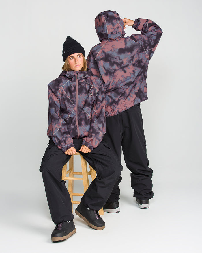 Two people in black and purple tie-dye Volcom Unisex Liftline 20K Jackets and black pants pose in a studio. One wears the baggy-fit snowboard jacket, sitting on a stool looking left, while the other stands with their back turned, hood up and hands raised to their head, showcasing the bold style and 20K waterproof performance of these Volcom jackets.