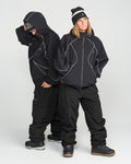 Two people in black winter outfits with white piping stand side by side. One faces forward with hands in pockets, wearing a beanie, while the other looks down, adjusting their hood. Both wear Volcom Unisex Liftline 20K Pants and snow boots—perfect for staying dry and comfortable on the slopes.
