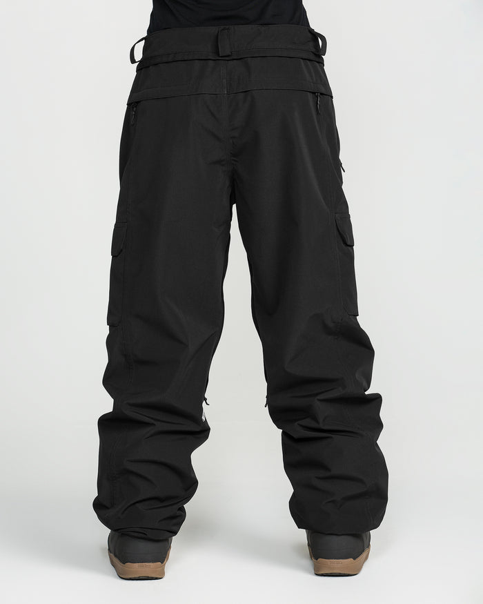 A person wearing loose-fitting black Volcom Unisex Liftline 20K Pants, made from recycled ski pant fabric, paired with brown boots and standing against a plain white background.