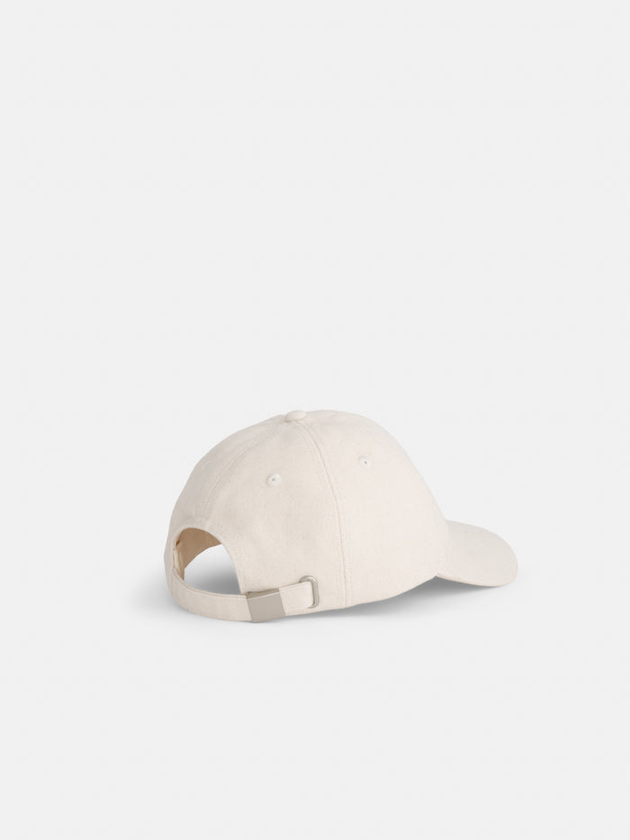 The Washed Canvas Baseball Cap is shown from the back at an angle, featuring a plain off-white design, a curved brim, and an adjustable strap with metal buckle.