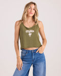 A woman with long blonde hair wears the Volcom Women's 91 Tank, featuring a graphic design and text, paired with blue jeans. She poses against a plain light background with one hand in her pocket.