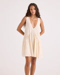 A woman with long, wavy hair stands against a white background, wearing the Volcom Women's A-Line Swing Dress. This cream-colored sleeveless mini dress is crafted from a soft viscose-cotton blend and features a deep V-neckline and gathered skirt. She poses with her arms behind her back, looking at the camera.