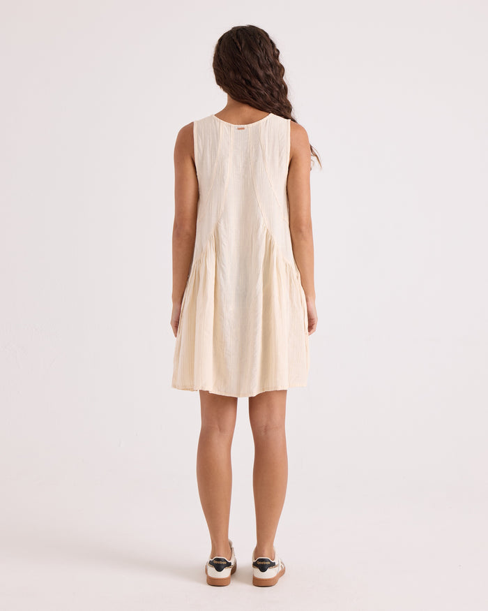 A woman with long, wavy hair stands facing away, wearing the Volcom Women's A-Line Swing Dress—a sleeveless mini dress in light beige, crafted from a soft viscose-cotton blend with a loose fit and pleated skirt. She is on a plain white background and pairs the dress with white sneakers featuring black details.