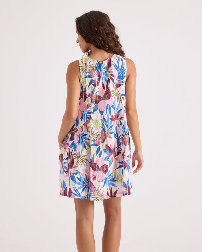 A woman stands facing away, wearing the Volcom Women's A-Line Swing Dress, which features a sleeveless design and a vibrant tropical leaf print in blue, pink, and beige hues. The flattering A-line silhouette is highlighted against the plain white background.
