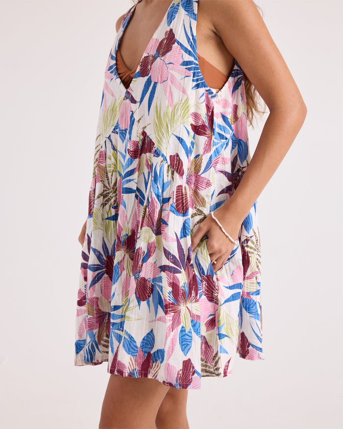 A woman models the Volcom Women's A-Line Swing Dress, a sleeveless mini dress featuring a loose, A-line silhouette and a colorful floral print in blue, pink, green, and burgundy. She has her hand in one of the pockets of this woven day dress and wears a beaded bracelet as an accessory.