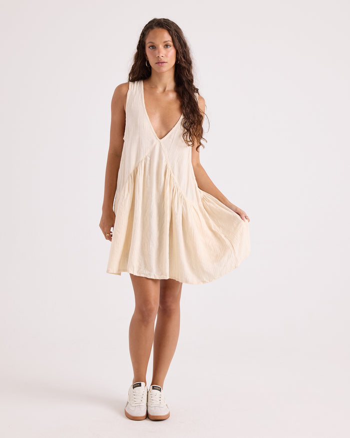 A woman wears the Volcom Women's A-Line Swing Dress—a cream-colored, sleeveless mini dress made from a soft viscose-cotton blend, featuring a V-neckline and a relaxed, flowing A-line skirt—paired with white sneakers and set against a plain white background.