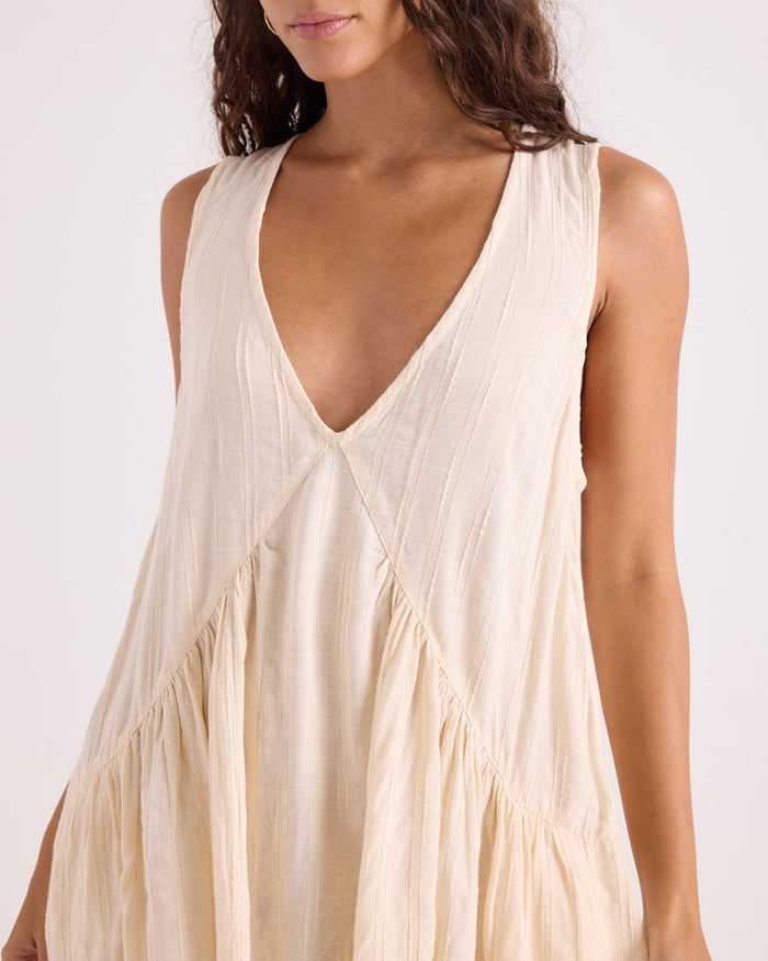 A woman models the Volcom Women's A-Line Swing Dress, a sleeveless, cream-colored dress featuring a deep V-neckline and gathered, flowing fabric. Crafted from a soft viscose-cotton blend, her face is partially out of frame against a plain white background.