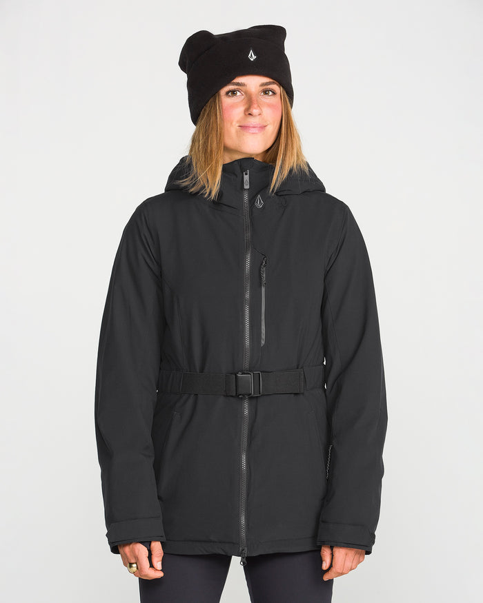 A woman wearing the Volcom Women’s Alpine All Day Insulated Jacket with a belt and a black beanie stands facing forward against a plain white background, smiling slightly.
