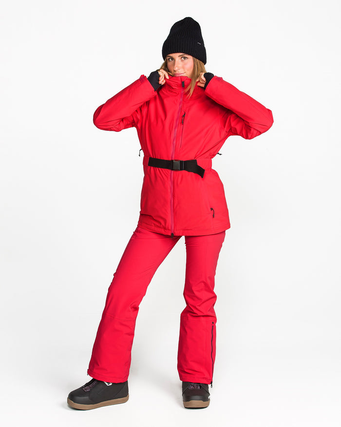 A person in a red Volcom Women’s Alpine All Day Insulated Jacket featuring 4-way stretch and 10K/10K V-Science technology, with matching pants, a black winter hat, and black boots stands against a white background, holding the jacket collar with both hands.