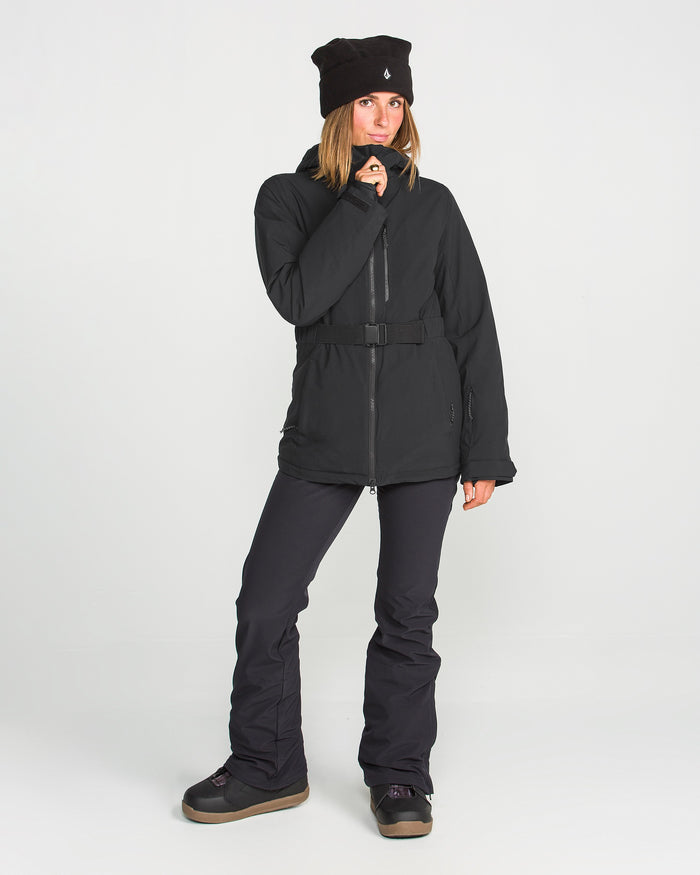 A woman stands against a white background, wearing the Volcom Women’s Alpine All Day Insulated Jacket in black, paired with black pants, a black beanie, and dark boots. She holds the jacket collar with one hand and gazes slightly to the side.