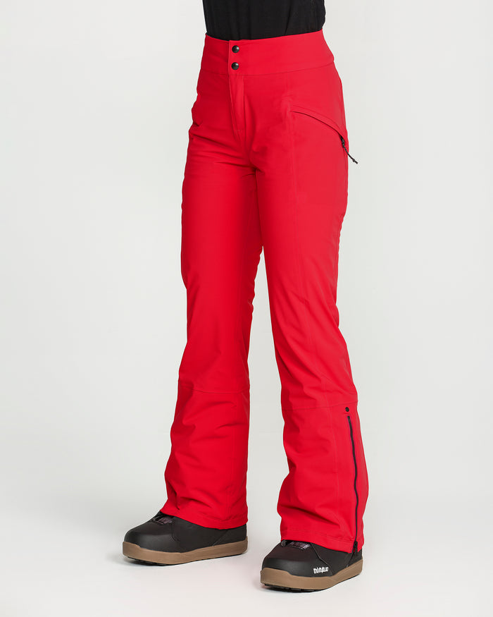 A person is wearing bright red Volcom Women’s Alpine All Day Insulated Pants and black snow boots, standing against a plain white background. These high-rise fit pants feature zippered pockets, flared legs, and two visible buttons at the waist, and are crafted with 4-way ultra stretch fabric for all-day comfort on or off the slopes.