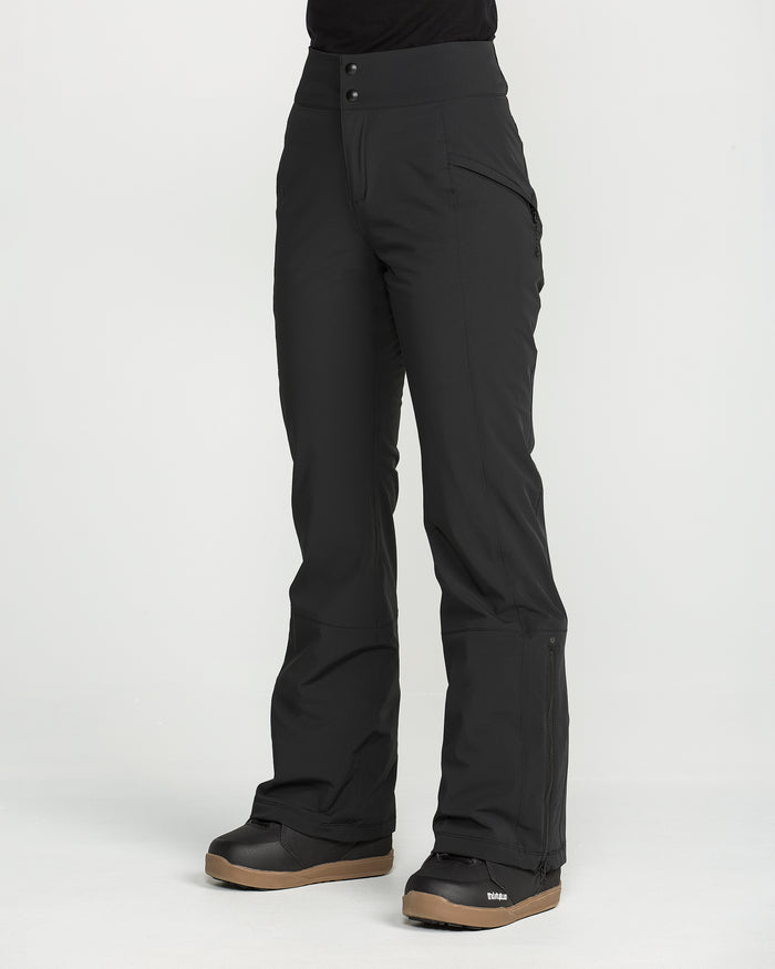 A person is shown wearing Volcom Women’s Alpine All Day Insulated Pants in black, featuring a high-rise fit and flared legs, paired with black snow boots with tan soles. The individual is standing on a white background, and only the lower body is visible.