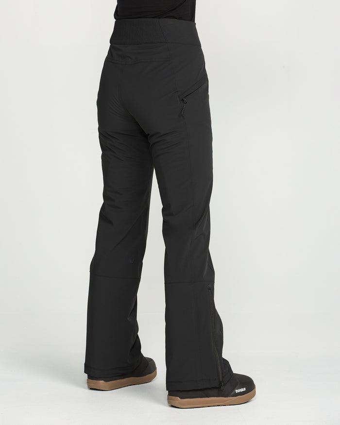A person is wearing the Volcom Women’s Alpine All Day Insulated Pants in black and black boots, standing on a white background and facing away from the camera. These pants feature a slim, high-rise fit with a zipped pocket and boot-cut hem.