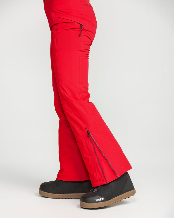 A person wearing Volcom Women’s Alpine All Day Insulated Pants in bright red, featuring 4-way ultra stretch and zippered side vents, paired with black snow boots. Posed sideways against a plain white background, only the lower body is visible.