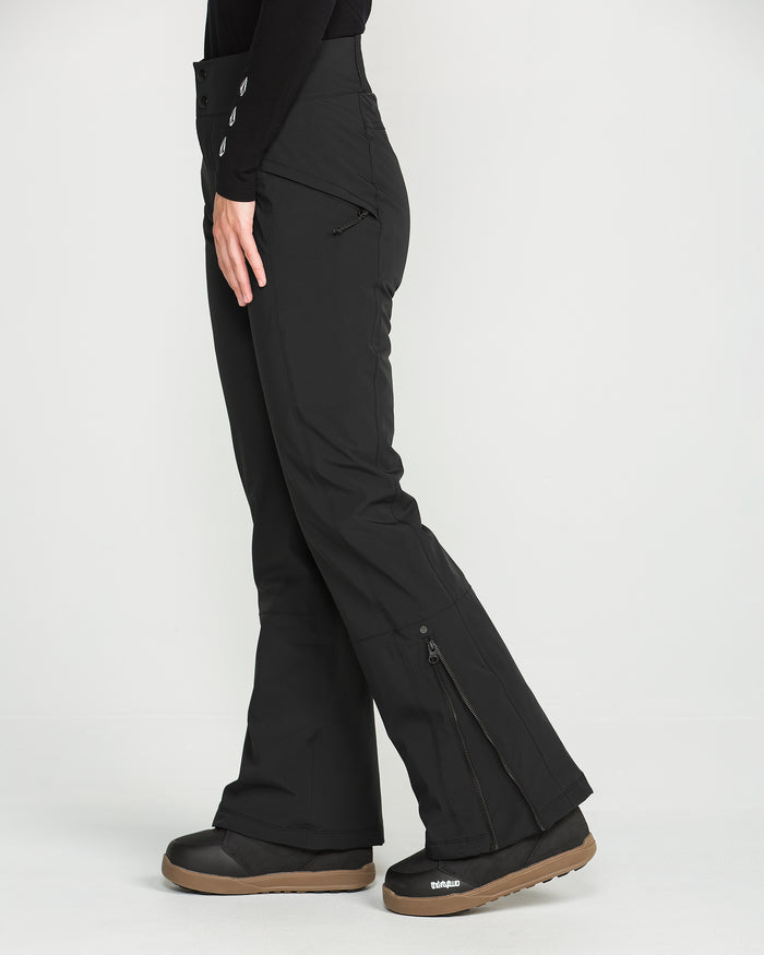 A person wearing black Volcom Women’s Alpine All Day Insulated Pants and black boots stands sideways against a plain white background. The pants, designed with a high-rise fit, feature zippers at the sides and a fitted waist.