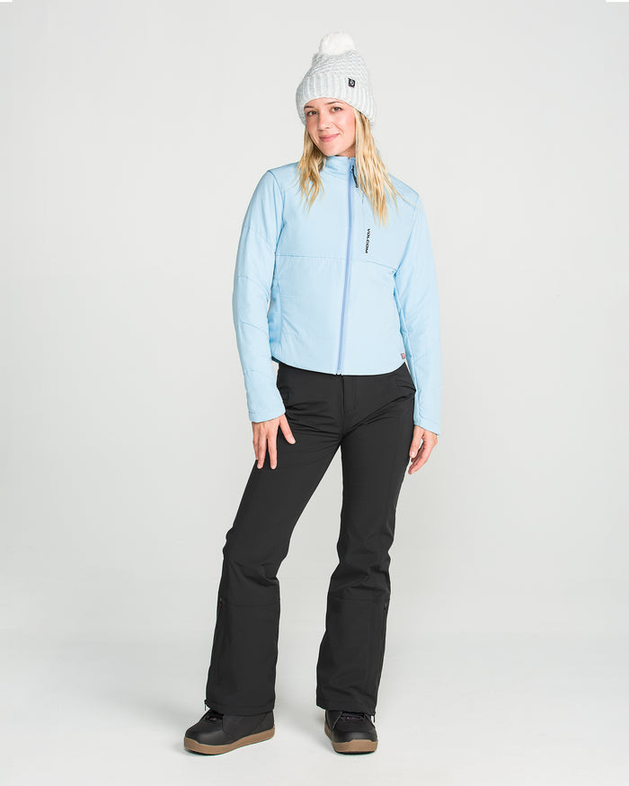 A woman wearing a light blue zip-up jacket, Volcom Women’s Alpine All Day Insulated Pants in black, black boots, and a gray knit hat with a white pom-pom stands smiling against a plain light background.