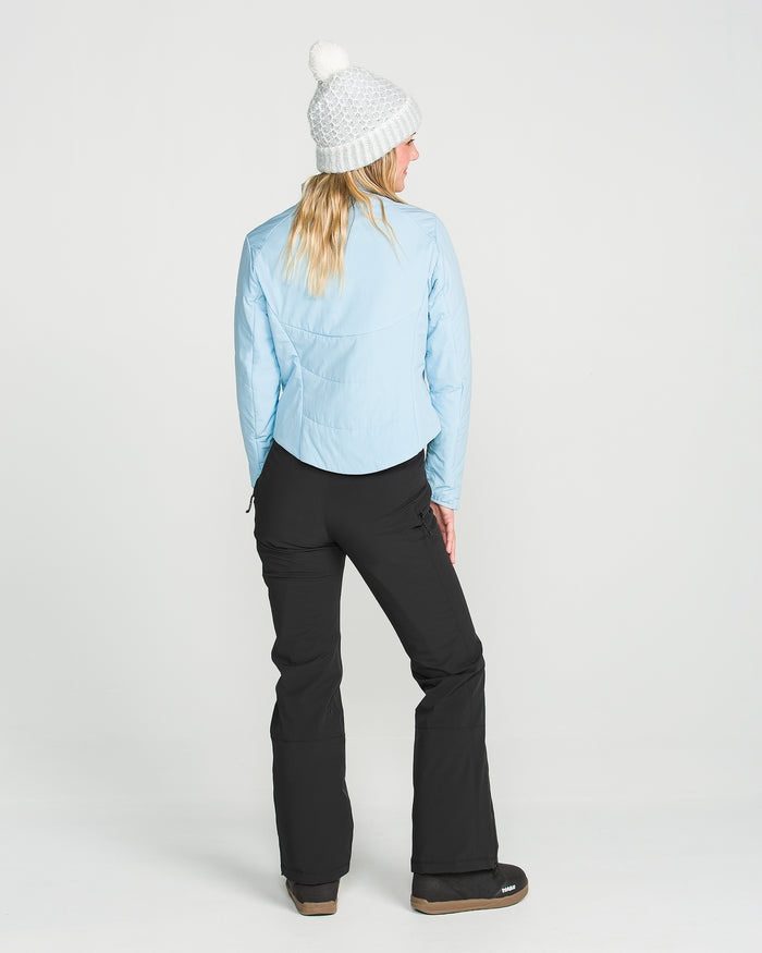 A woman wearing a white knit hat, light blue jacket, and Volcom Women’s Alpine All Day Insulated Pants with a high-rise fit stands facing away, looking over her left shoulder in a studio with a plain white background. The pants feature recycled insulation for enhanced comfort and sustainability.